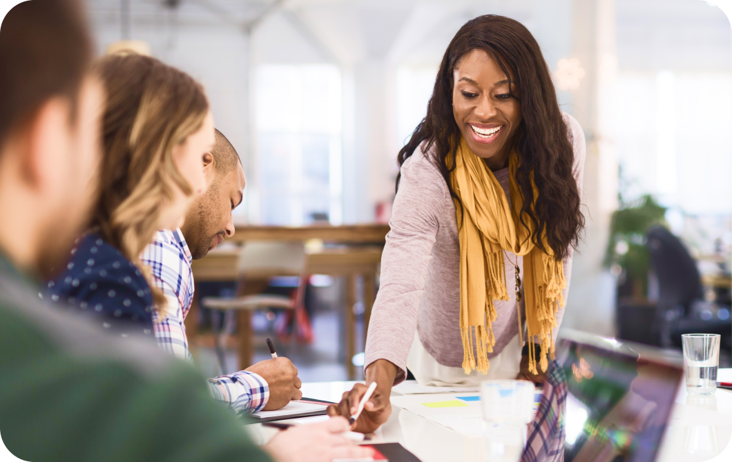 A smiling professional black woman points to a significant part of a document while her coworkers take notes.