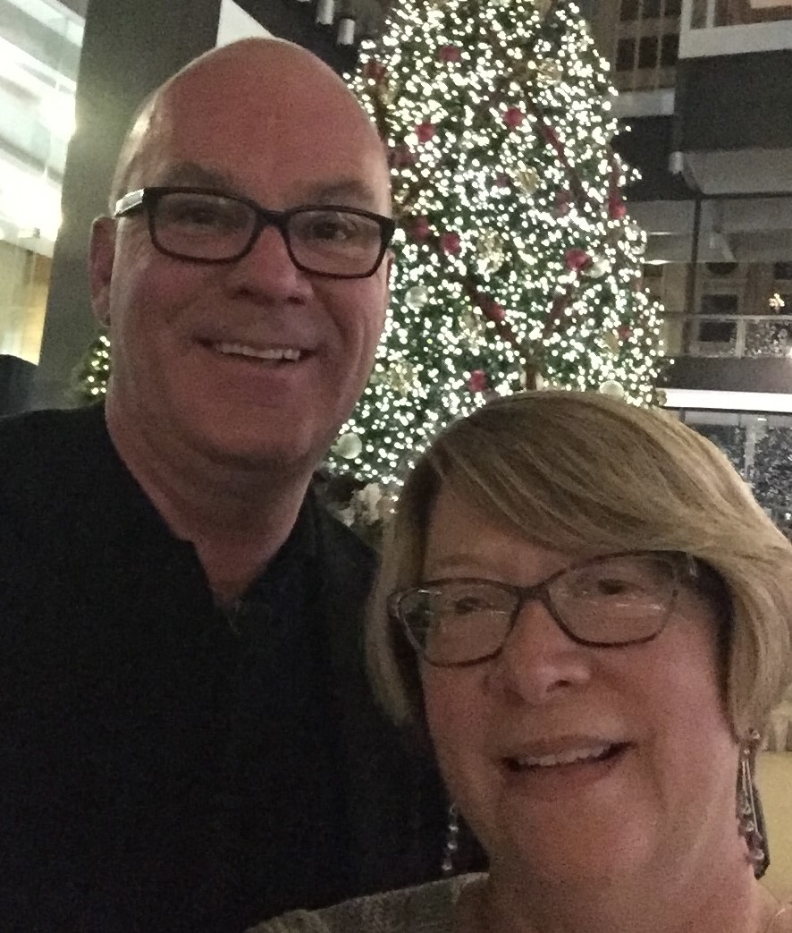 A smiling man and woman stand in front of a lit up Christmas tree.