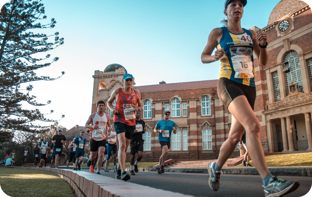 Runners wearing race bibs run on a path on a university campus.