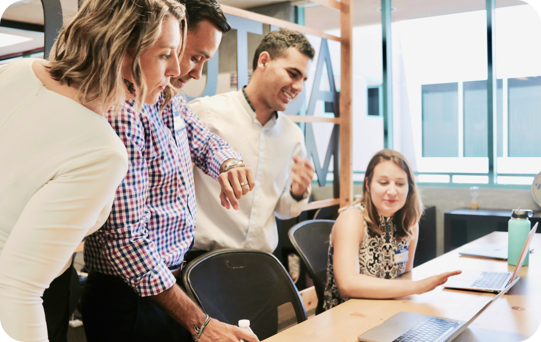 unsplash_e6n7uoEnYbA A seated woman with a laptop in front her gestures as three standing colleagues lean over the table.