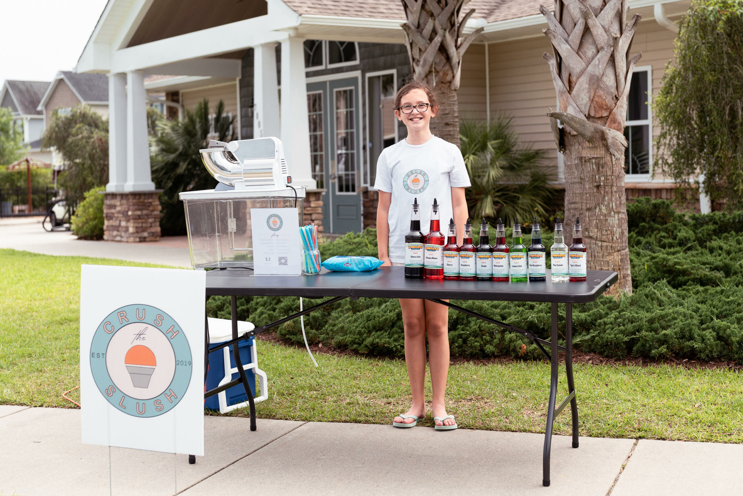 A smiling young girl stands behind a table loaded up with slushie making supplies.
