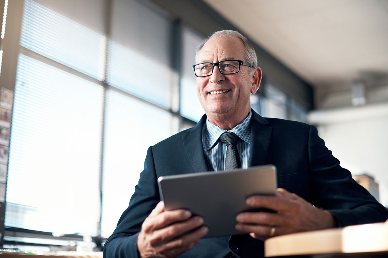 An older smiling man in a suit holds a tablet in front of his chest.