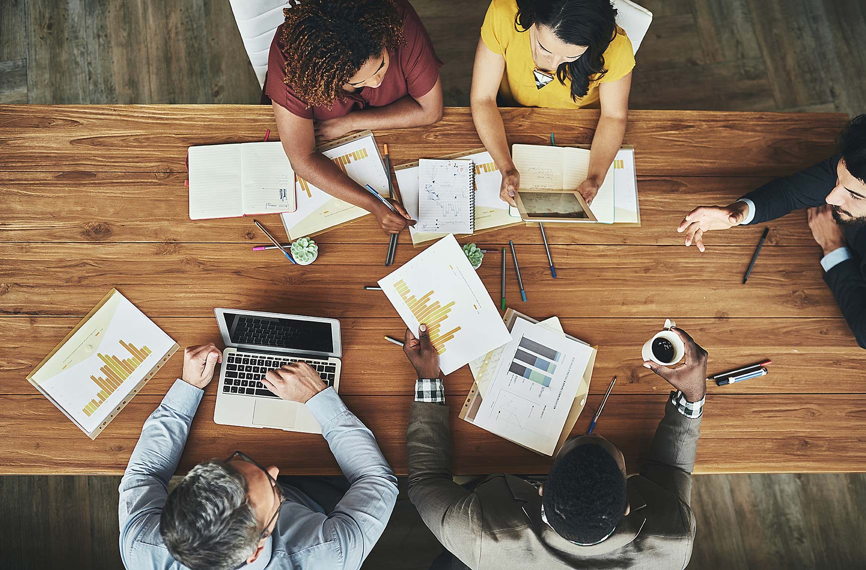 Top view of a group of five people sitting around a table with papers, a laptop and a tablet spread out in front of them.