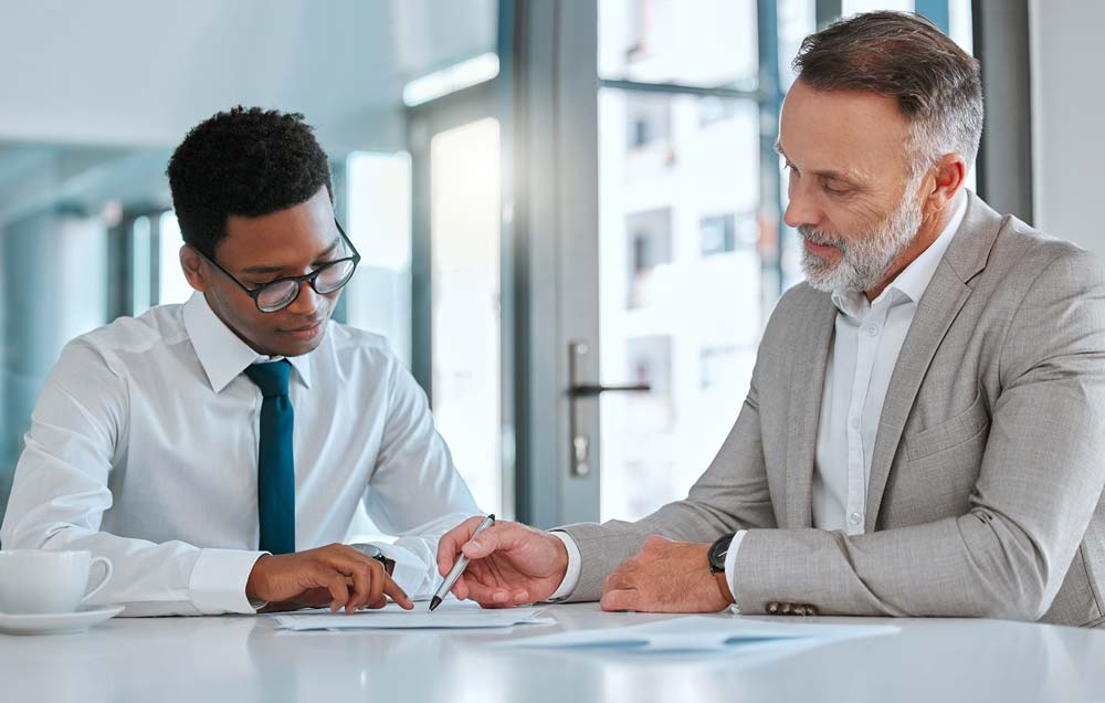 A younger professional black man wearing glasses and an older white man in a suit look at something on the paper in front of them.