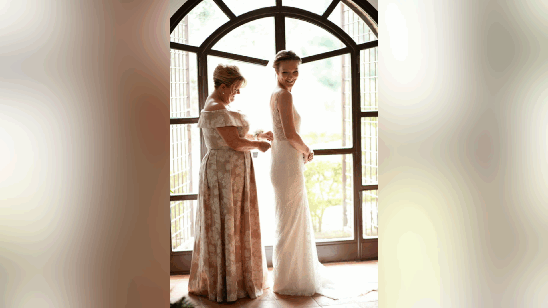 A joyful moment between a mother and daughter as they prepare for the wedding, with the mother helping fasten the bride’s dress in front of a bright arched window.