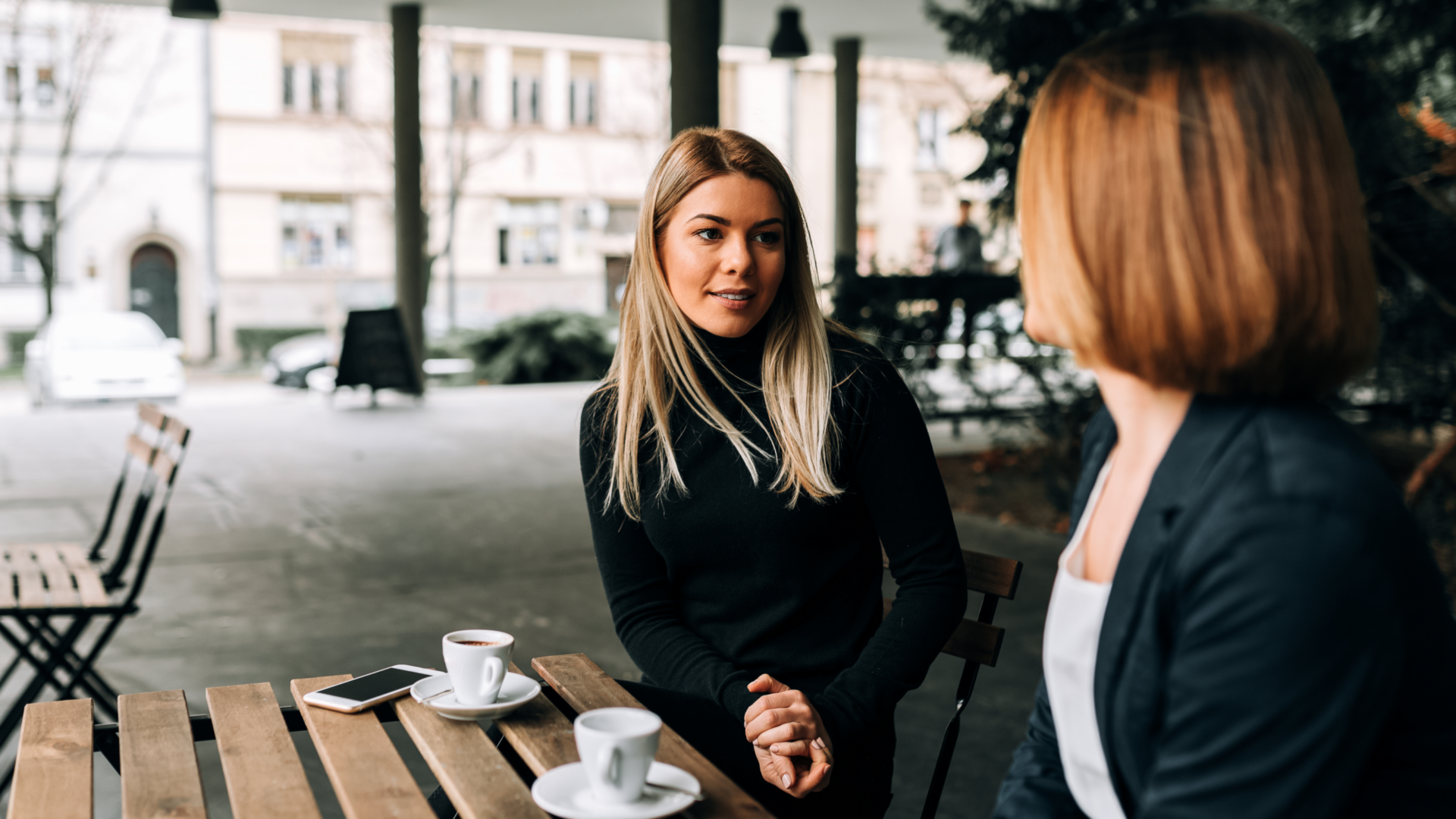 Two women seated at an outdoor café table engaged in conversation over coffee, leaning in attentively in a warm, relationship focused setting.