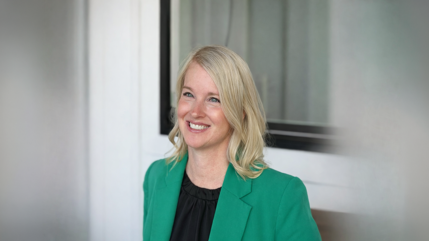 A smiling woman with shoulder-length blonde hair wears a green blazer over a black blouse, seated outdoors against a softly blurred background with a window behind her.