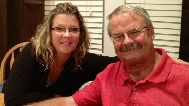 A woman and an older man sit closely together at a table, smiling warmly at the camera in a cozy indoor setting.