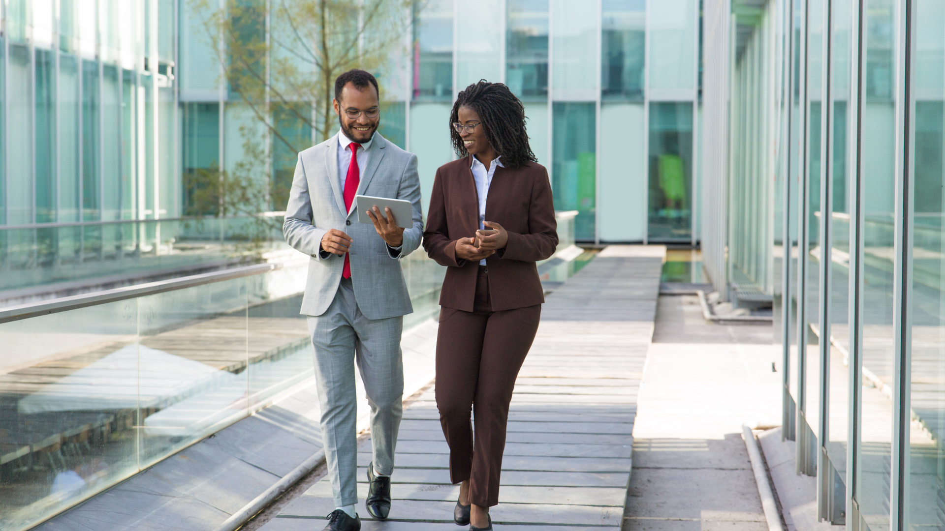 Two business professionals walk together through a modern office courtyard, smiling and discussing information displayed on a tablet.
